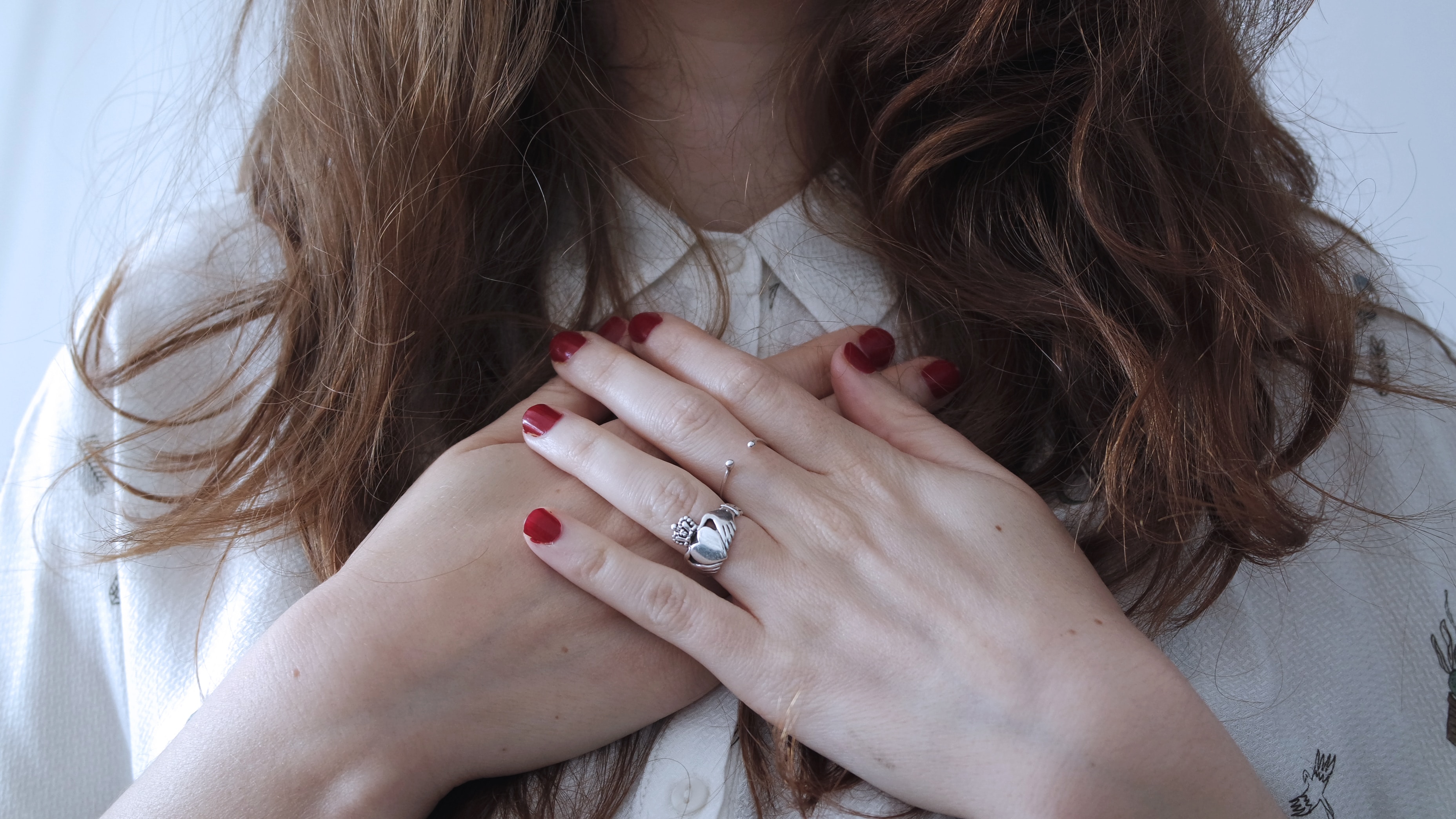 a person with long hair, red nails, and rings, holding their hands over the center of their chest